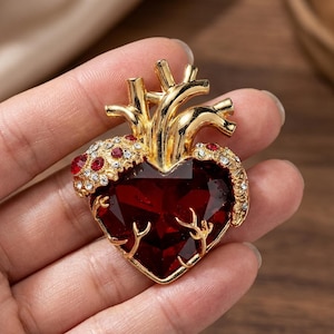 Puede incluir: Broche dorado en forma de corazón, con una gran piedra preciosa roja oscura facetada en el centro. El broche tiene vetas doradas y una parte superior dorada con detalles de cristal rojo y transparente.