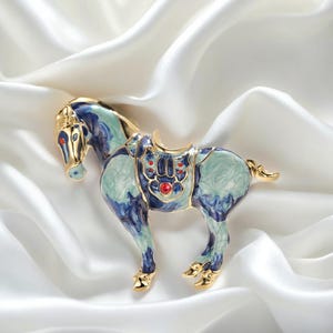 Pode incluir: Broche de cavalo dourado com detalhes em esmalte azul e verde-azulado. O cavalo tem uma sela com detalhes vermelhos, sobre um fundo branco e sedoso. Acessório decorativo.