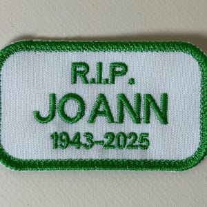 Puede incluir: Un parche rectangular blanco con un borde verde. El parche tiene el texto "R.I.P. JOANN 1943-2025" bordado en verde.