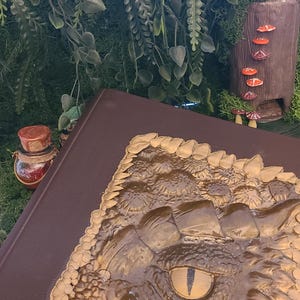 May include: A brown book with a detailed, gold-colored embossed design of a reptile eye and scales. The book is set against a backdrop of greenery and a miniature woodland scene with mushrooms and a small bottle.