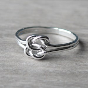 May include: A silver-colored ring with a knot design. The ring features a double band and a detailed knot centerpiece, showcasing a polished, reflective surface. The ring is set against a neutral background, highlighting its intricate design.