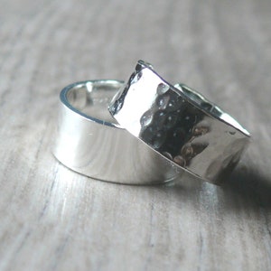 May include: Two silver rings. One ring has a smooth, polished surface, while the other has a hammered texture. The rings are displayed on a light-colored wooden surface. The rings are likely for a wedding or engagement.