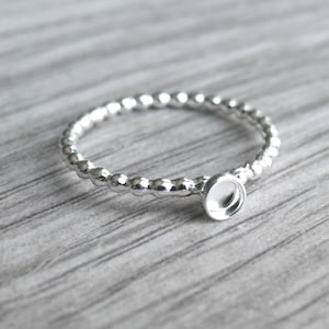 May include: A silver-colored ring with a beaded band and a small, round setting. The ring is designed to hold a small stone or embellishment. The ring is shown on a light gray wooden surface.