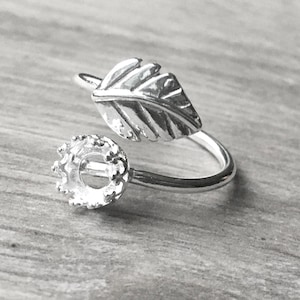 May include: A silver-colored ring with a leaf design and a setting for a gemstone. The leaf is detailed with veining, and the setting is round with prongs. The ring is on a wooden surface.