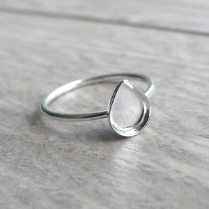 May include: A silver ring with a teardrop-shaped setting. The ring has a thin band and a bezel setting designed to hold a gemstone or other decorative element. The ring is shown on a light gray wooden surface.
