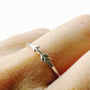 May include: A delicate silver ring featuring an arrow design. The ring is thin and sits on a finger, showcasing the intricate arrow detailing. The ring is a simple yet elegant piece of jewelry.