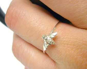 May include: A silver ring featuring a detailed bee design. The bee has textured wings and body, and is attached to a thin, delicate band. The ring is shown on a finger, highlighting the intricate details of the jewelry.
