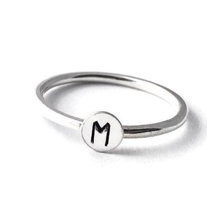 Pode incluir: Um anel prateado com uma banda fina e um berloque circular. O berloque apresenta a letra "M" em preto, centrada em um fundo branco. O anel est&aacute; sobre um fundo branco.