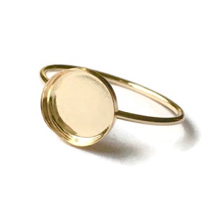 May include: A gold-colored ring with a round bezel setting. The ring band is thin and delicate, and the bezel is designed to hold a round stone or cabochon. The ring is set against a white background.
