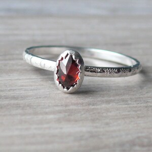 May include: A silver ring with a faceted, oval-shaped, deep red gemstone. The ring band has a textured, hammered appearance. The gemstone is set in a decorative bezel setting, creating a classic and elegant jewelry piece.