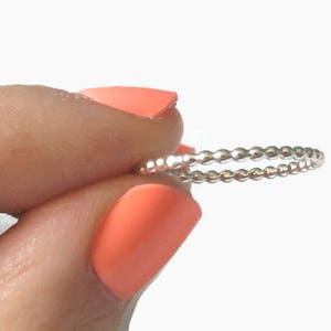 May include: A delicate silver ring featuring a beaded design. The ring is held between fingers with orange nail polish, set against a white background. The ring's design consists of small, evenly spaced beads.