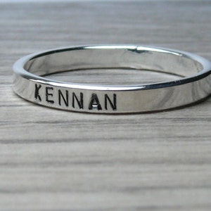 May include: A silver ring with the name "KENNAN" engraved on the outside. The ring is a thin band, likely made of sterling silver, and sits on a light-coloured wooden surface.
