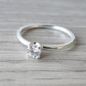 Puede incluir: Un anillo de plata con una sola gema redonda y transparente. La banda del anillo es delgada y pulida, y la gema est&aacute; engastada en un engaste de cuatro puntas. El anillo se muestra sobre una superficie de madera gris claro.