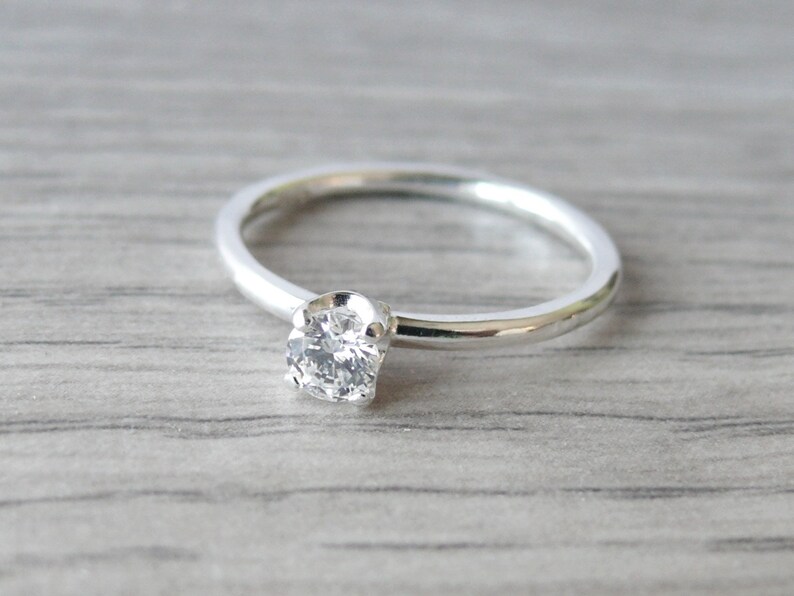 Puede incluir: Un anillo de plata con una sola gema redonda y transparente. La banda del anillo es delgada y pulida, y la gema est&aacute; engastada en un engaste de cuatro puntas. El anillo se muestra sobre una superficie de madera gris claro.