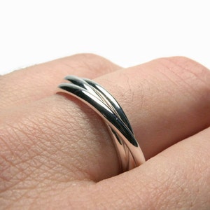May include: A close-up of a silver ring with a unique design. The ring features two curved bands that intertwine, creating a flowing, elegant look. The polished metal reflects light, highlighting the smooth, rounded surfaces. The ring is worn on a finger.