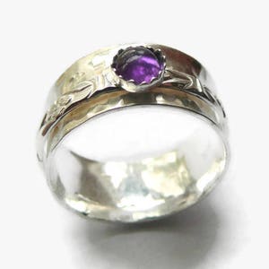 May include: A silver ring with a hammered texture and a bezel-set, round, purple gemstone. The ring features a floral design on the band. The ring is a wide band style.