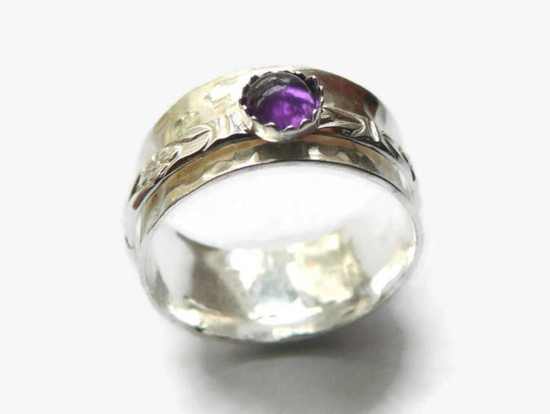 May include: A silver ring with a hammered texture and a bezel-set, round, purple gemstone. The ring features a floral design on the band. The ring is a wide band style.