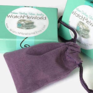 Pode incluir: Duas caixas turquesa com o texto "WatchMeWorld" e uma imagem de anel na frente. Uma bolsa roxa com cord&atilde;o est&aacute; em frente &agrave;s caixas. As caixas s&atilde;o de cor verde-azulado claro.