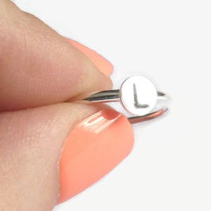 Pode incluir: Um anel de prata com uma face circular com a letra "L". O anel &eacute; segurado entre os dedos com esmalte de unhas cor de coral. A banda do anel &eacute; fina e a letra est&aacute; centrada na face.