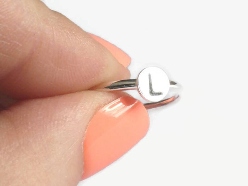 Pode incluir: Um anel de prata com uma face circular com a letra "L". O anel &eacute; segurado entre os dedos com esmalte de unhas cor de coral. A banda do anel &eacute; fina e a letra est&aacute; centrada na face.