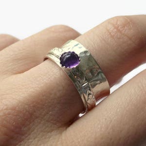 May include: A silver ring with a hammered texture and a floral design. The ring features a round, deep purple gemstone. The ring is worn on a finger.