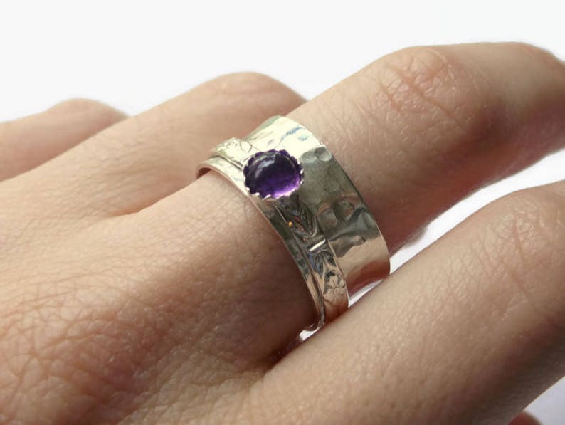 May include: A silver ring with a hammered texture and a floral design. The ring features a round, deep purple gemstone. The ring is worn on a finger.