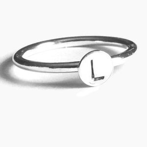 Pode incluir: Um anel de prata com uma face circular com a letra "L". A banda do anel &eacute; fina e polida, e a letra est&aacute; centrada na face. O anel est&aacute; sobre um fundo branco.