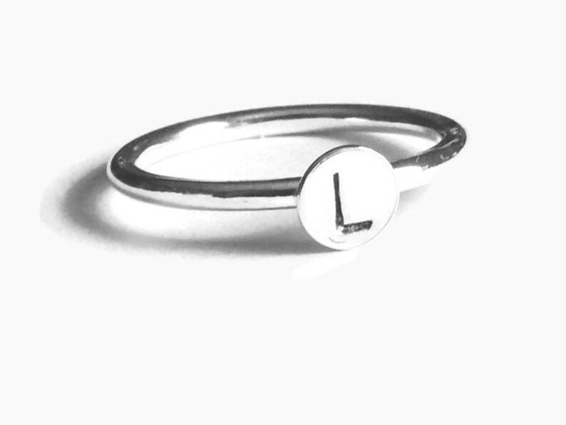 Pode incluir: Um anel de prata com uma face circular com a letra "L". A banda do anel &eacute; fina e polida, e a letra est&aacute; centrada na face. O anel est&aacute; sobre um fundo branco.
