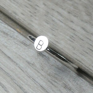Pode incluir: Um objeto de metal prateado com um pequeno detalhe circular branco com a letra "B". Est&aacute; sobre uma superf&iacute;cie texturizada cinza claro, imitando o gr&atilde;o da madeira. O design &eacute; simples e moderno.