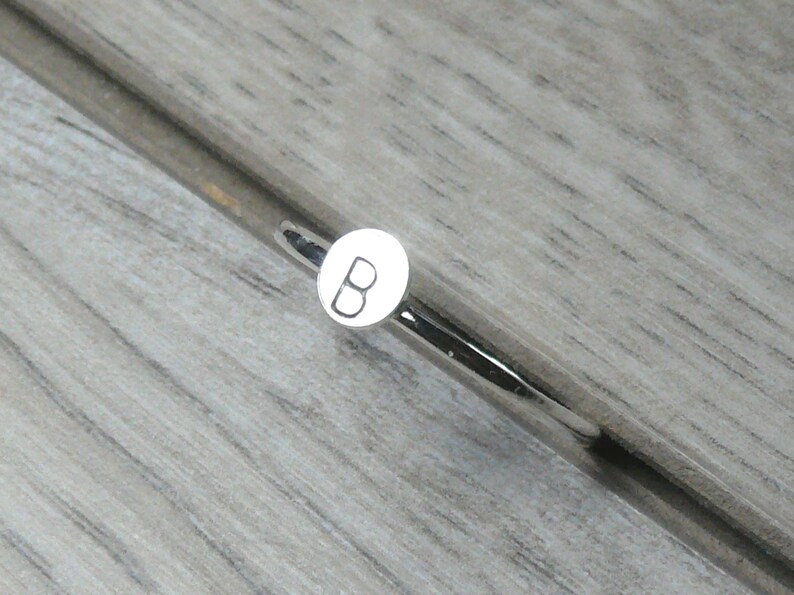 Pode incluir: Um objeto de metal prateado com um pequeno detalhe circular branco com a letra "B". Est&aacute; sobre uma superf&iacute;cie texturizada cinza claro, imitando o gr&atilde;o da madeira. O design &eacute; simples e moderno.