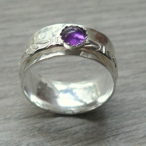 May include: A silver ring with a wide band and a hammered texture. The ring features a bezel-set, round, purple gemstone. The band has a floral design. A piece of jewelry.