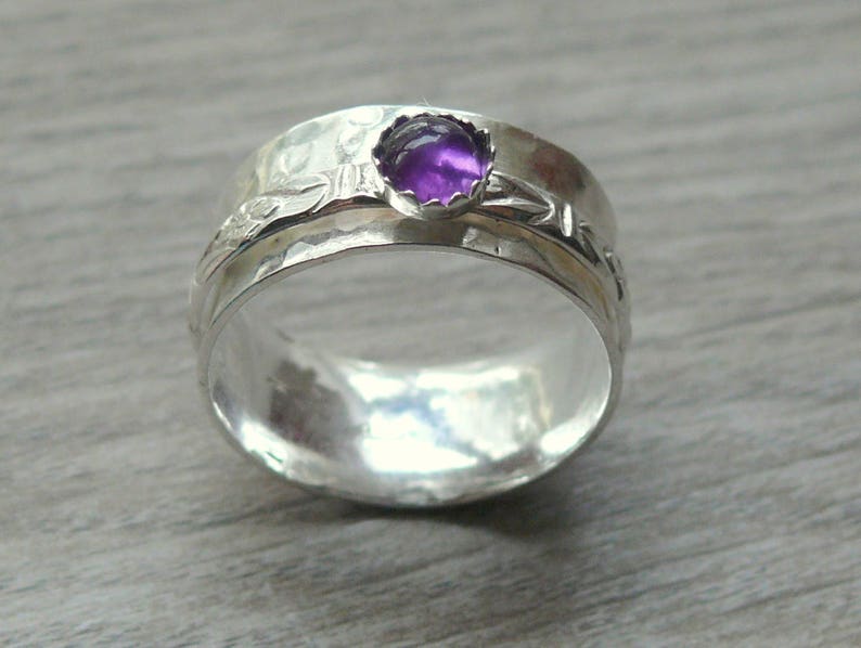 May include: A silver ring with a wide band and a hammered texture. The ring features a bezel-set, round, purple gemstone. The band has a floral design. A piece of jewelry.