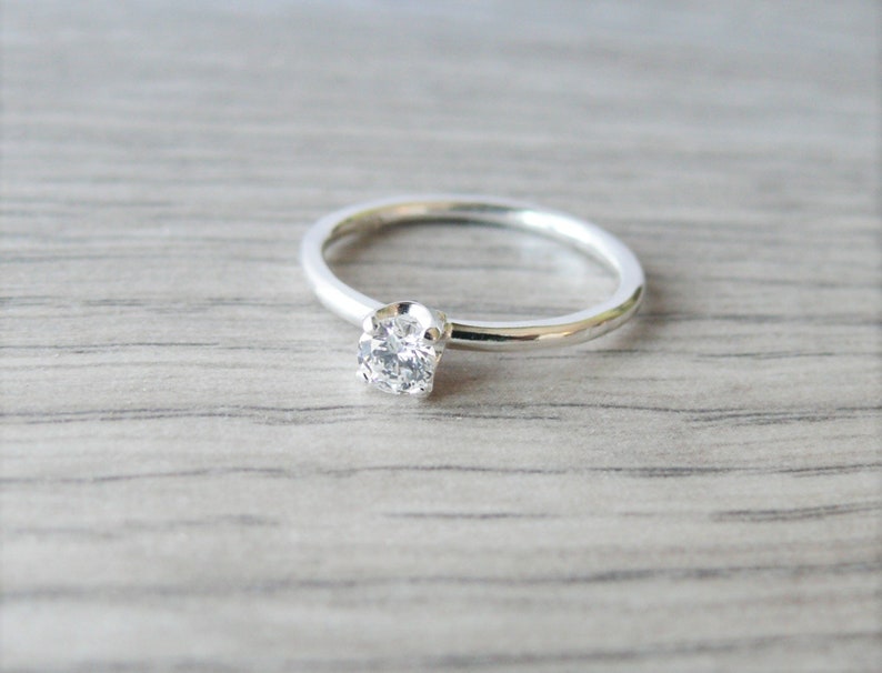 Puede incluir: Un anillo de plata con una sola gema redonda y transparente. La banda del anillo es delgada y pulida, y la gema est&aacute; engastada en un engaste de cuatro puntas. El anillo se muestra sobre una superficie de madera gris claro.