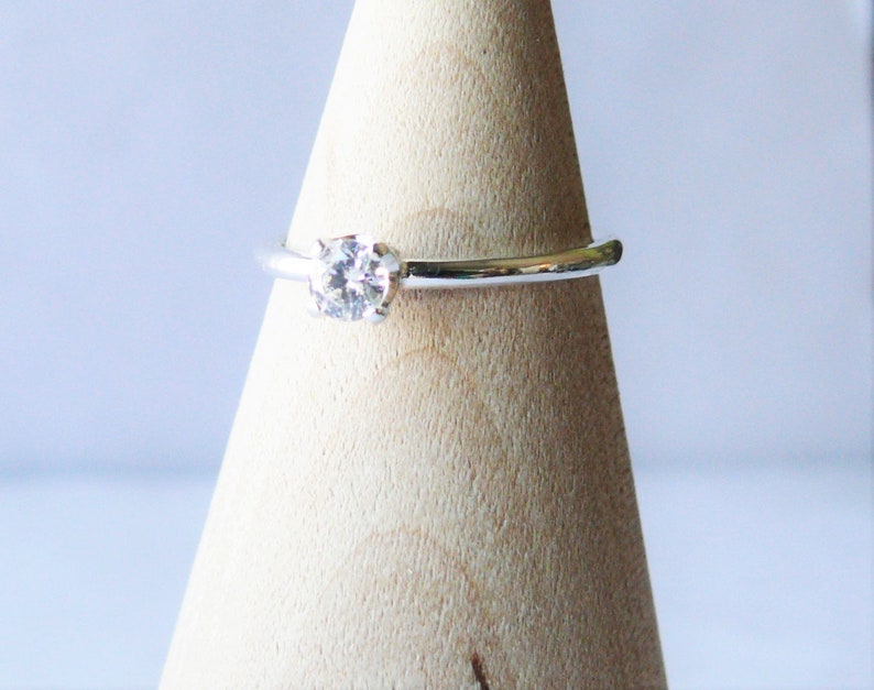 Puede incluir: Un anillo de plata con una sola gema redonda y transparente. El anillo est&aacute; engarzado en una banda sencilla y se muestra sobre un soporte de madera en forma de cono. La gema brilla, captando la luz.