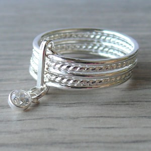 May include: A silver ring with multiple bands, some with a twisted rope design. A small, clear gemstone hangs from the ring. The ring is displayed on a light-colored wooden surface. Jewelry.