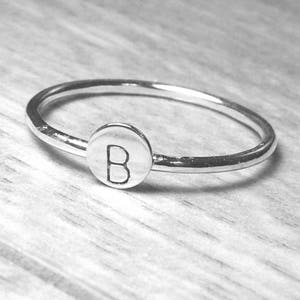 Pode incluir: Um anel de prata com um berloque circular com a letra "B". O anel est&aacute; sobre uma superf&iacute;cie de madeira clara. A banda do anel &eacute; fina e polida, com o berloque centrado.