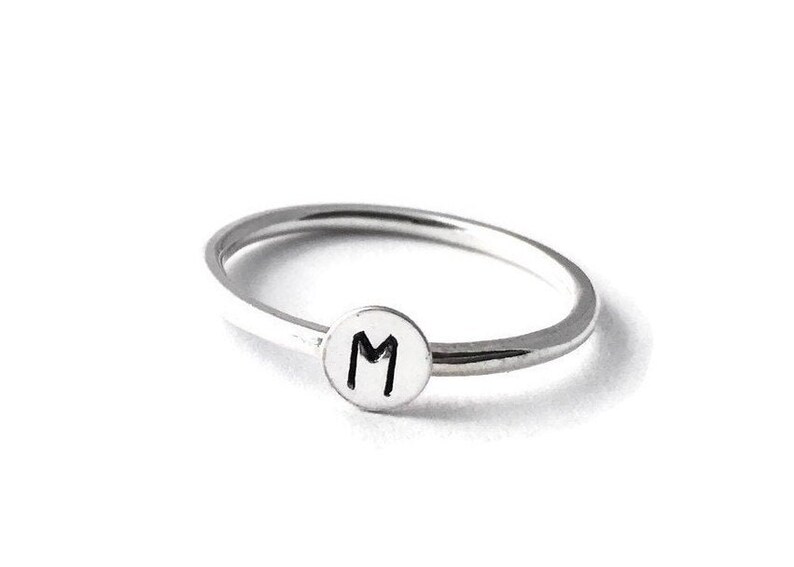 Pode incluir: Um anel prateado com uma banda fina e um berloque circular. O berloque apresenta a letra "M" em preto, centrada em um fundo branco. O anel est&aacute; sobre um fundo branco.
