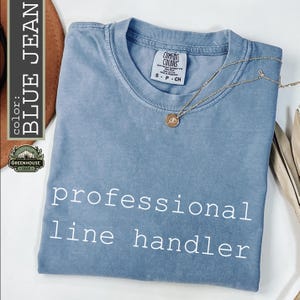 May include: A blue jean colored short-sleeve t-shirt with a crew neckline. The shirt features the white text 'professional line handler' printed on the front. A gold-toned necklace with a small circular pendant rests on the fabric near the collar.