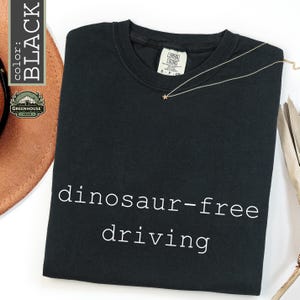 May include: A black short-sleeved t-shirt with a crew neckline featuring the white text "dinosaur-free driving" printed on the front. The shirt has a Comfort Colors brand label on the inside collar and a thin gold-toned necklace resting on it.
