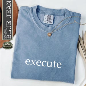 May include: A blue short-sleeved t-shirt with a crew neckline and the word execute printed in white on the front. The shirt features a Comfort Colors label at the inner neck and is paired with a thin gold-toned chain necklace with a small round pendant.