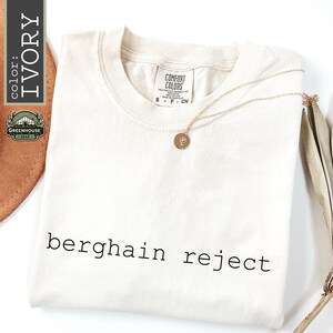 May include: An ivory short-sleeved t-shirt with a crew neckline featuring the black text "berghain reject" printed on the front. The shirt has a Comfort Colors brand label inside the collar and is paired with a thin gold chain necklace with a small round pendant.