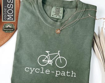 May include: A moss green t-shirt with a white bicycle graphic and the words "cycle-path" printed below. A gold necklace with a pendant rests on the shirt. The shirt is from Greenhouse Gear.