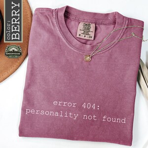 May include: A berry-colored short-sleeve t-shirt with a crew neckline. The shirt features the white text "error 404: personality not found" printed on the front. A gold-toned necklace with a small circular pendant rests on the fabric.