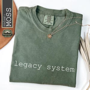 May include: A moss green crew neck t-shirt featuring the text legacy system printed in white across the chest. The shirt has a Comfort Colors brand label inside the collar and is displayed with a gold-toned necklace resting on the fabric.