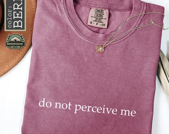 Peut inclure: Un t-shirt couleur baie avec une encolure ronde et le texte blanc "do not perceive me" imprimé sur le devant. Le t-shirt présente une étiquette de marque Comfort Colors à l'intérieur du col, indiquant la taille S, et un collier doré fin.