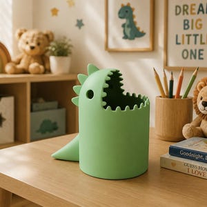 May include: A light green dinosaur-shaped desk tidy with an open mouth design. The organiser is on a wooden table, with a plush lion, pencils, and books nearby. The background includes framed artwork and a wooden shelf with toys.