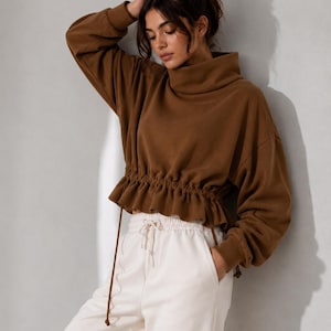 Könnte beinhalten: Ein braunes Langarm-Sweatshirt mit hohem Stehkragen, überschnittenen Schultern und einer gerafften Taille mit Kordelzug, die einen Rüschensaum bildet, kombiniert mit einer cremeweißen Jogginghose mit elastischem Kordelzugbund.