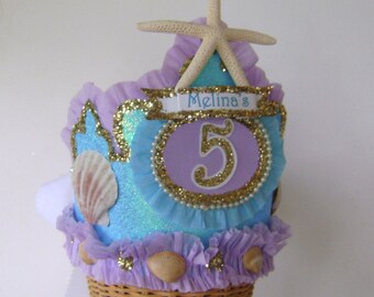 May include: A purple and blue birthday crown with a starfish on top, a number 5, and the name "Melina's" on a banner. The crown is decorated with seashells and glitter.
