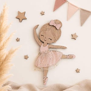 Peut inclure: Une décoration murale en bois représentant une ballerine avec un corsage peint en rose, une jupe en tulle à paillettes dorées et un nœud rose assorti. L'ensemble comprend plusieurs petites étoiles en bois et une guirlande de trois fanions.
