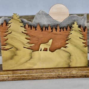 May include: A wooden landscape scene featuring a howling wolf silhouette, trees, mountains, and a sun. The artwork is crafted from various wood tones and mounted on a rectangular base, creating a three-dimensional effect.